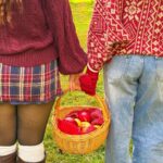 Two people in autumn sweaters and a plaid skirt holding a basket of apples.