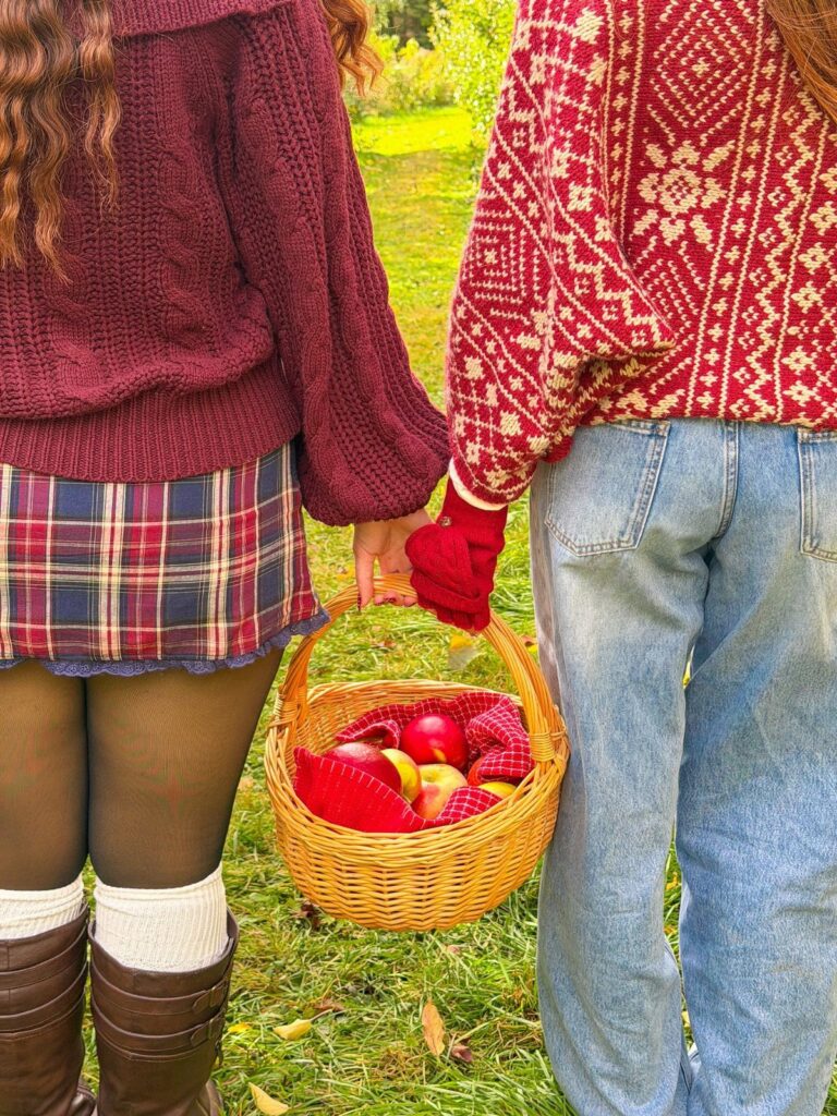 Two people in autumn sweaters and a plaid skirt holding a basket of apples.