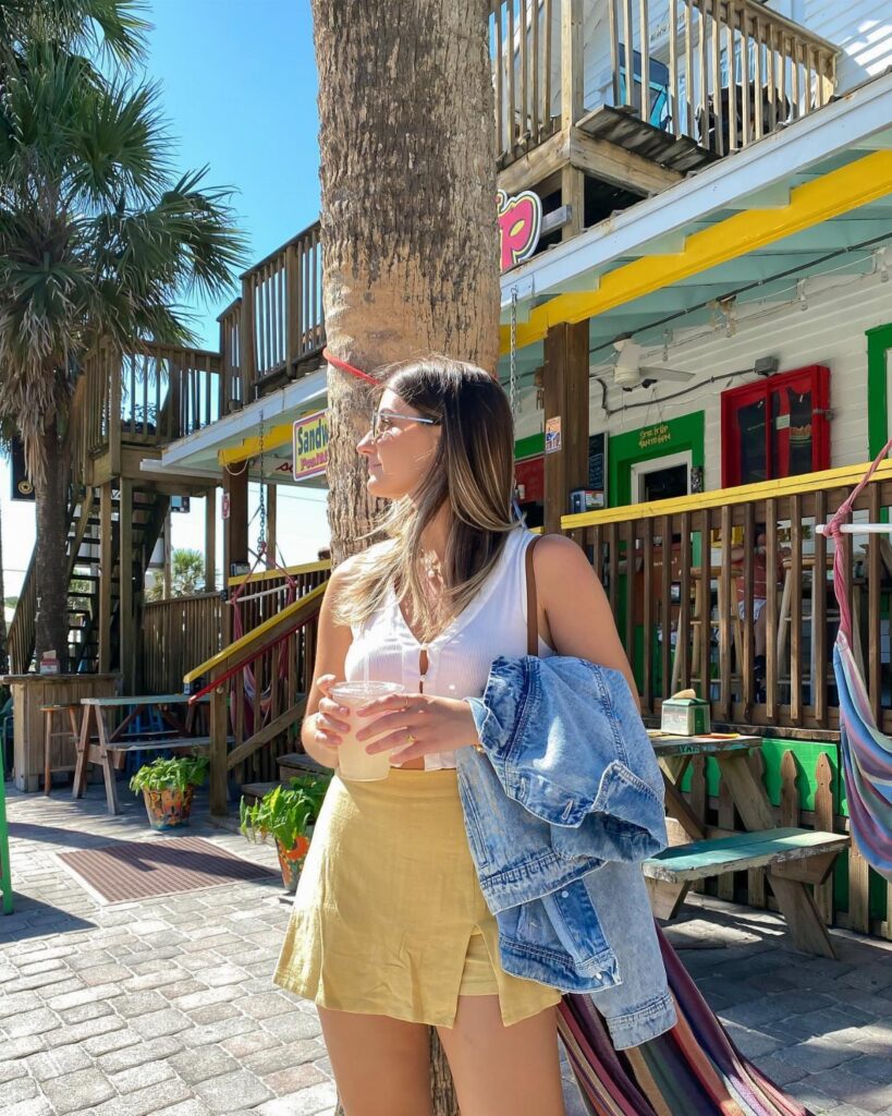 A woman in a white top and yellow skirt standing by a tree near a beachside building.