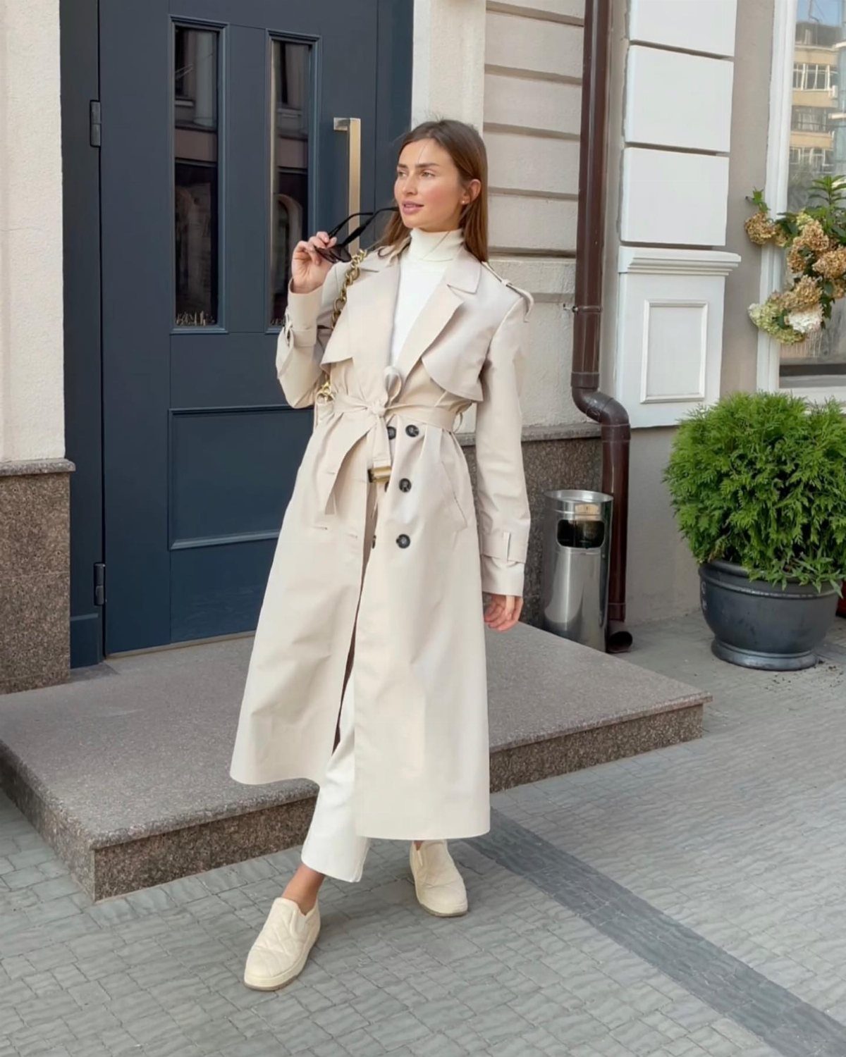 Woman in a long beige trench coat and white trousers standing outdoors.