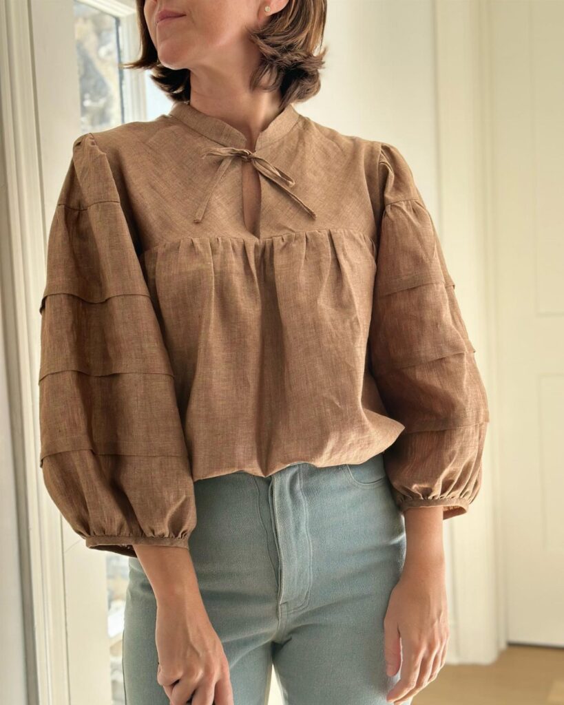 Woman wearing a brown linen peasant blouse with tiered puff sleeves and light blue jeans.