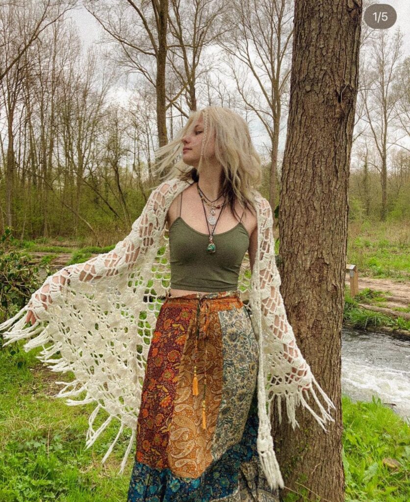 Woman in a green tank top, patterned skirt, and white crochet cardigan posing by a tree.