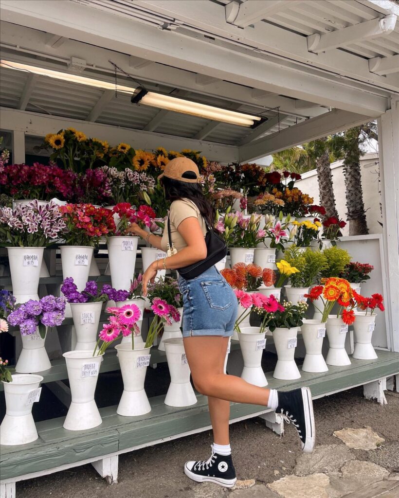 Woman in denim shorts and tank top posing by colorful flowers in white vases.