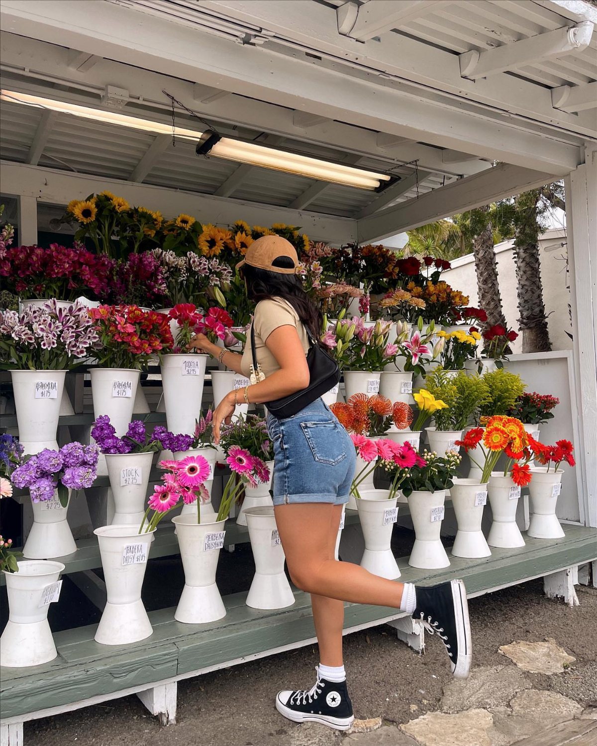 Woman in denim shorts and tank top posing by colorful flowers in white vases.