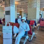 Woman in grey matching lounge set sitting in airport terminal with silver suitcase.