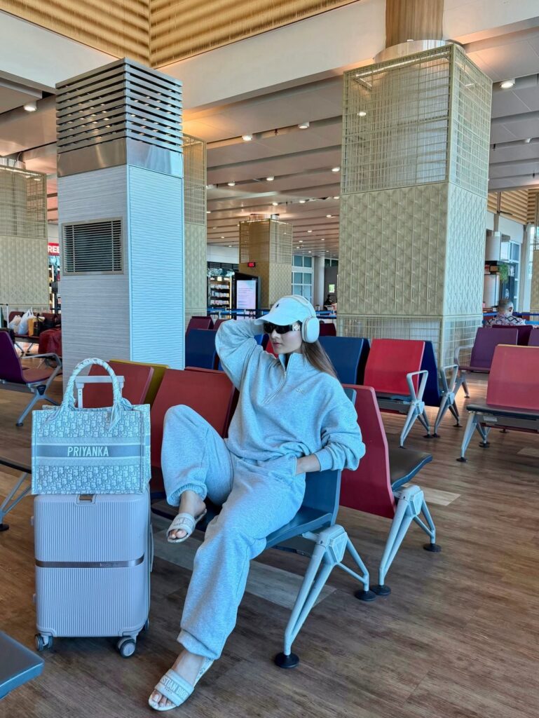 Woman in grey matching lounge set sitting in airport terminal with silver suitcase.