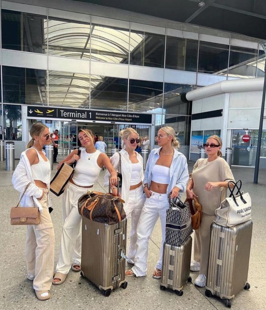 Five young women posing with suitcases in front of an airport terminal entrance.