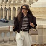 Woman in a brown suede jacket, striped shirt, and cream wide-leg trousers walking outdoors.