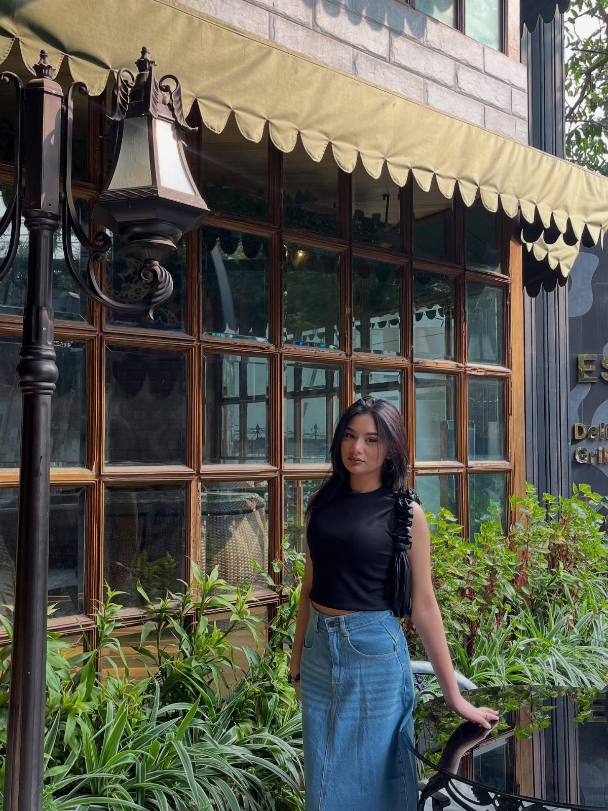 Woman wearing a black top and long blue denim skirt standing outdoors.
