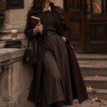 Woman in a long chocolate brown coat and plaid skirt standing outdoors in autumn.