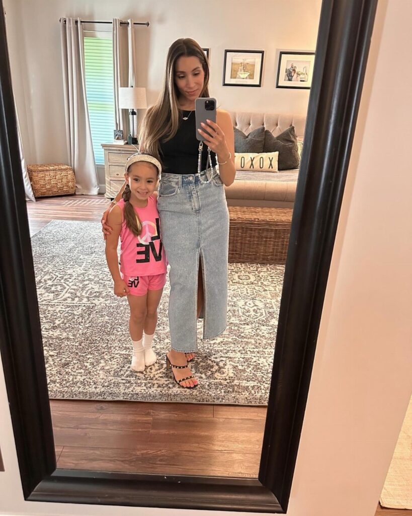 Woman in black tank and long denim skirt posing with daughter in pink outfit.