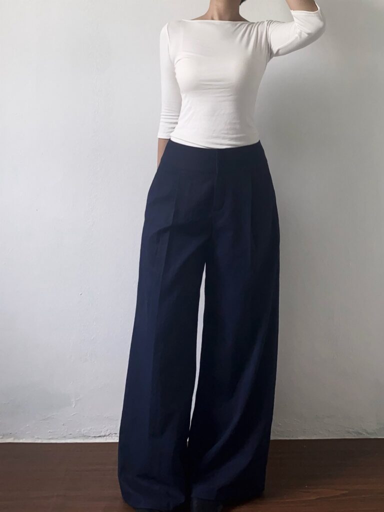 A person wearing a white long-sleeved shirt and navy blue wide-leg trousers against a white wall.