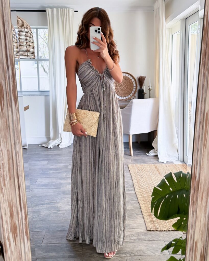 Woman in a long patterned strappy maxi dress holding a straw clutch in a mirror selfie.