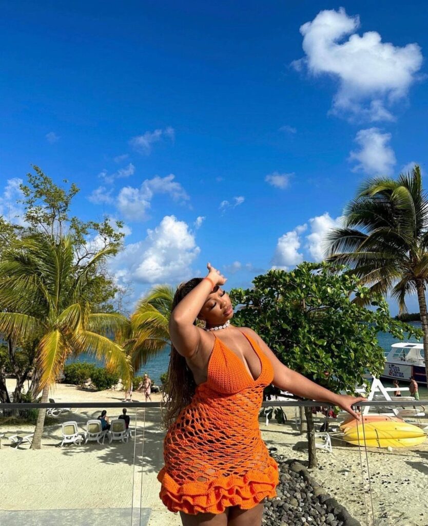 Woman in a bright orange crochet mini dress posing on a tropical beach.