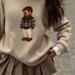 Beige bear sweater with white collared shirt and brown plaid pleated mini skirt.