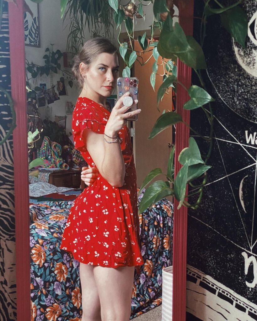 A young woman in a red floral summer dress posing for a mirror selfie in a room with plants.