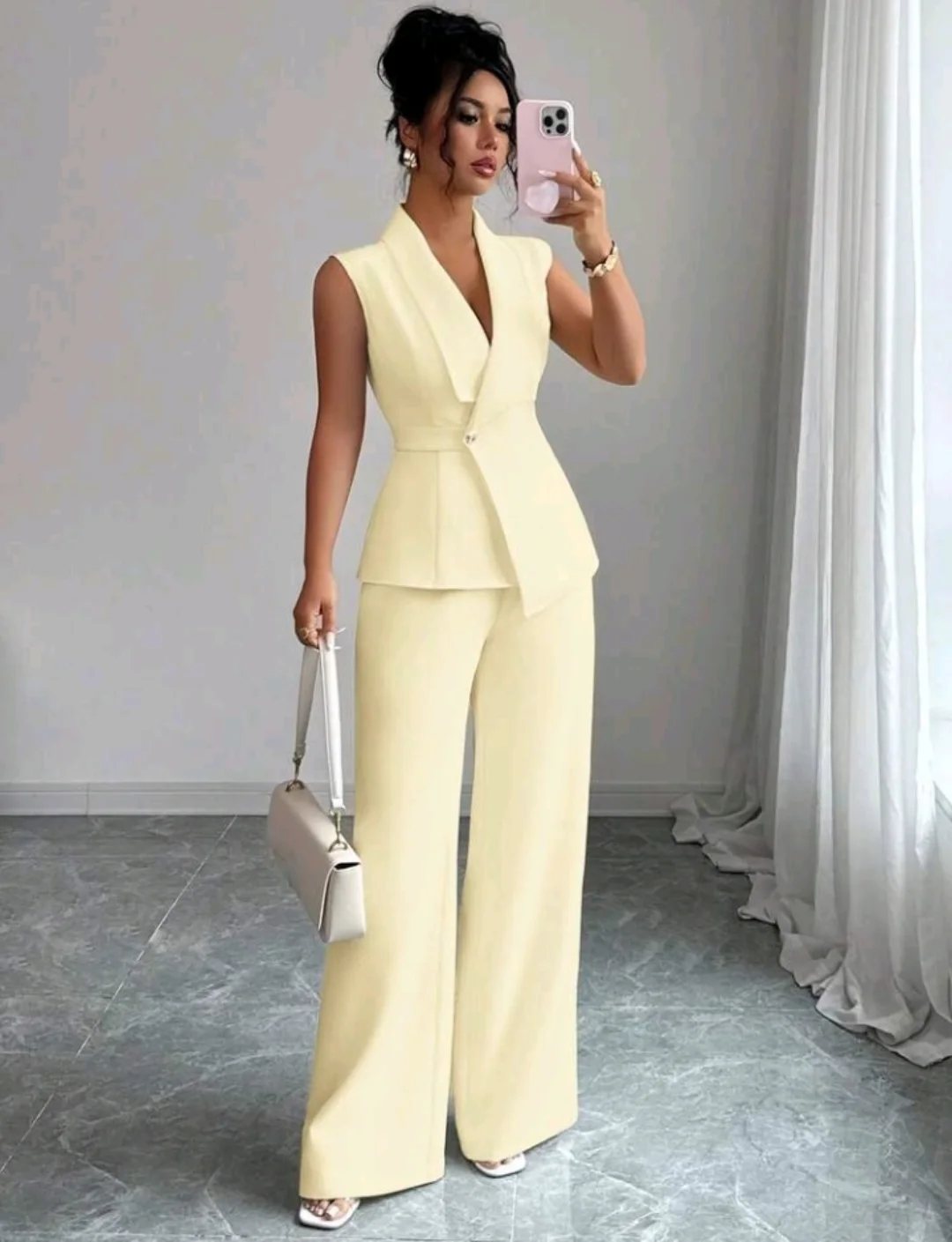 A woman wearing a pale yellow sleeveless blazer and matching wide-leg trousers while holding a white handbag.