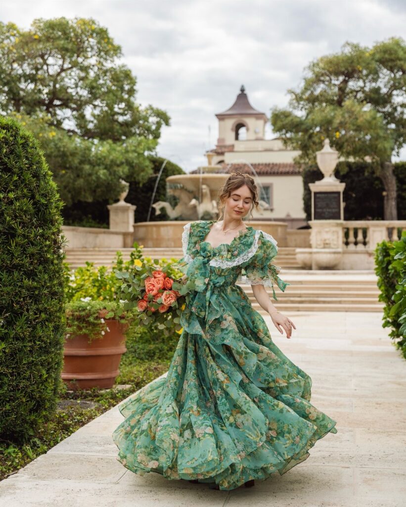 A woman wearing a green floral lace dress posing in a lush garden with a fountain.