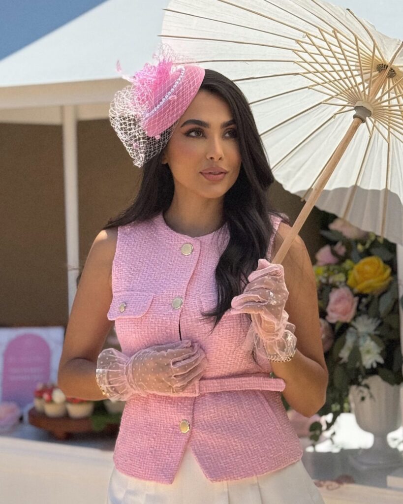 Woman in pink tweed vest and fascinator holding a white parasol outdoors.