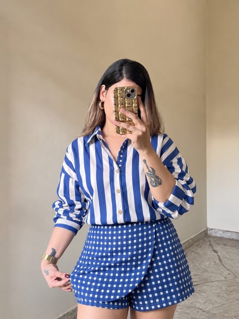 Woman in a blue and white striped shirt and gingham shorts posing for a mirror selfie.
