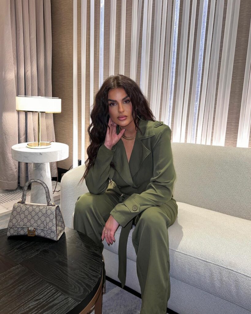 Woman in an olive green jumpsuit sitting on a light grey sofa in a room.