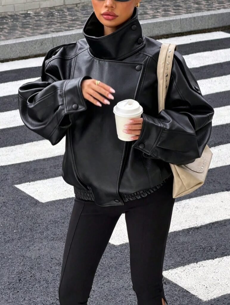 Woman in an oversized black leather jacket and black pants holding a coffee cup on a street.
