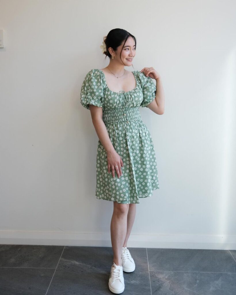 Woman wearing a green floral midi dress and white sneakers against a white wall.